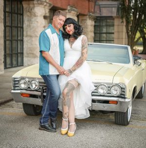 Two people pose together in front of a cream-colored vintage Chevrolet Impala with chrome bumpers and styled headlights.