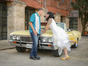 A vintage yellow convertible car parked on a street serves as a backdrop for a retro-styled wedding photo.