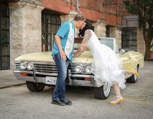A couple poses playfully in front of a vintage yellow Chevrolet classic car parked on an urban street.