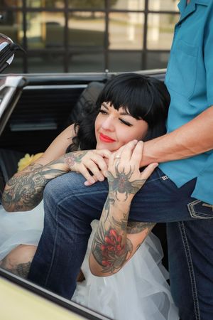 A bride in a white dress with tattoos and red lipstick sits in a car while holding someone's hand.