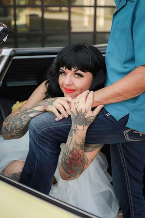 A woman with black bangs and red lipstick sits in a car wearing a white tulle dress, showing tattooed arms.