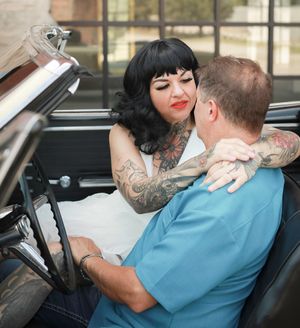 Couple in vintage convertible car as tattooed person in white dress embraces passenger.