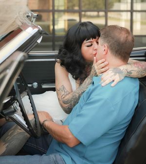 A couple shares an intimate moment while sitting in a vintage convertible car, with tattooed arms and retro styling visible.