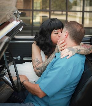 A romantic couple shares a kiss in a vintage convertible car, with the woman wearing a white dress and showing tattooed arms.