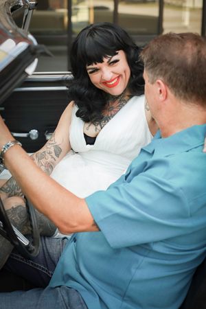 Two people sit in a convertible car, one wearing a white dress with dark hair and tattoos smiles at their companion.