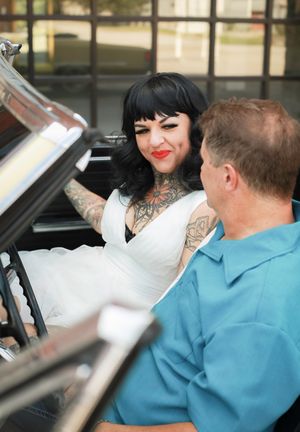 Couple sitting in vintage convertible car with the woman wearing white dress and displaying tattoo sleeves.