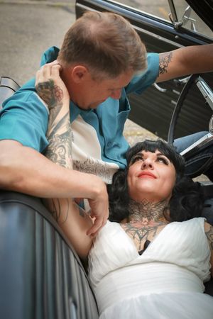 A tattooed bride in white dress reclines in a classic car as her partner leans in close.