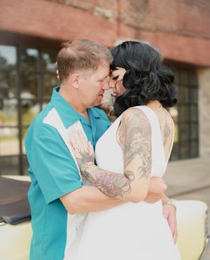 A romantic couple shares an intimate moment together, with one wearing a turquoise shirt and the other in a white dress with tattoos.