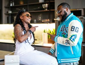 Two people having an animated conversation at a bar, one wearing a white tank top and another in a light blue varsity jacket.