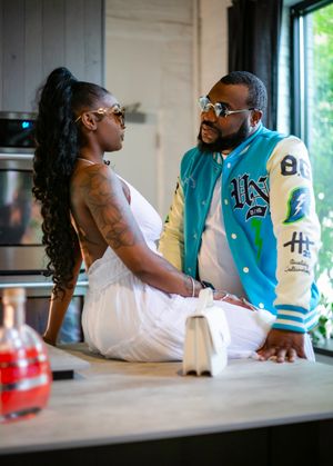 A stylish couple in sunglasses interact in a kitchen, with her in a white dress and him wearing a varsity jacket.