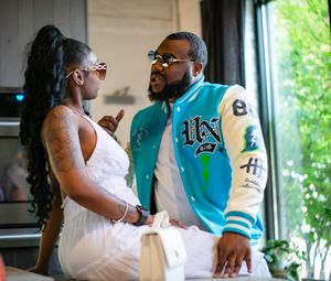 A couple shares a candid moment together indoors, with one wearing a white dress and the other in a turquoise varsity jacket.