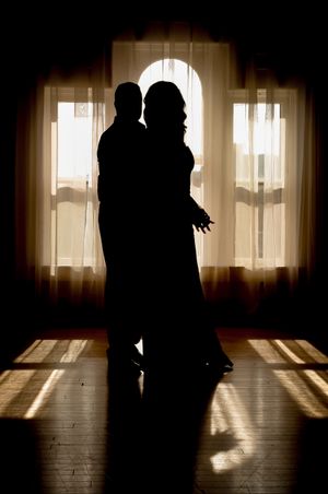 Silhouetted figures dance in front of sunlit curtained windows casting dramatic shadows on wooden floors.