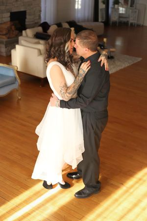 A couple shares their first dance on a sunlit hardwood floor in front of a cream-colored couch at their wedding.