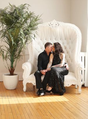 Romantic couple shares an intimate moment on an ornate white throne chair next to a potted palm plant on hardwood floors.