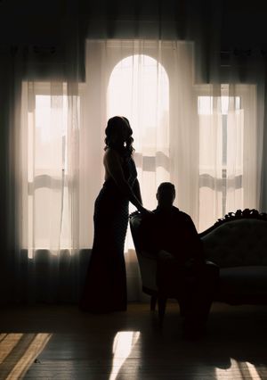Silhouetted figures stand in elegant pose by arched window with dramatic backlighting creating romantic ambiance.