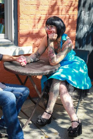 Person with tattoos wearing a blue dress and glasses sits at an outdoor cafe table against an orange brick wall.