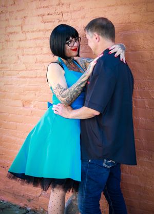 A couple in retro style clothing embraces against a brick wall, with the woman wearing a bright turquoise dress and glasses.