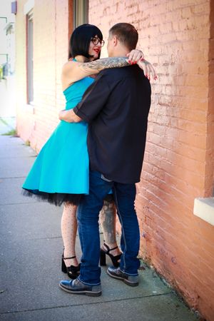A couple embraces against a brick wall, with her wearing a vibrant turquoise dress and black petticoat.