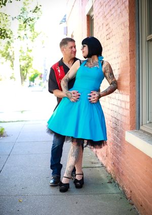 A couple embraces on a sidewalk against a brick wall, with the woman wearing a bright turquoise dress and showing tattoos.