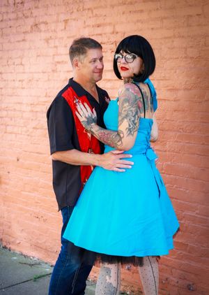 Rockabilly couple poses against brick wall with woman in bright blue dress and tattoos and man in black and red shirt.
