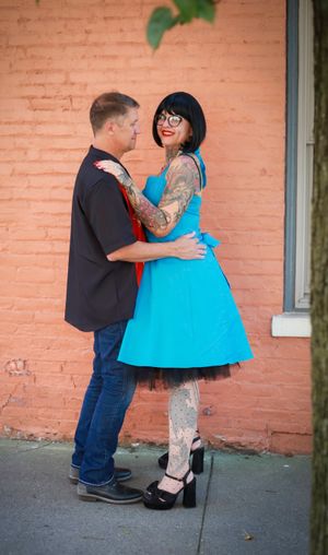 A couple poses together against an orange brick wall, with the woman wearing a bright turquoise vintage-style dress with tulle.