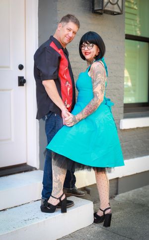 A couple in rockabilly style poses on steps, with a turquoise dress and tattoos against a light building exterior.