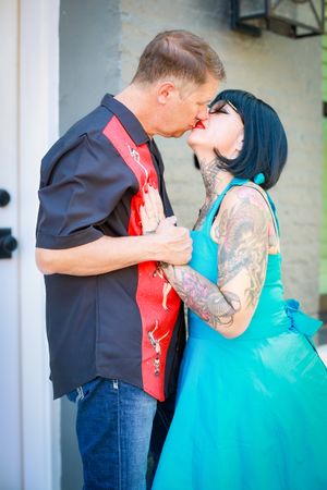 A romantic couple in rockabilly style clothing share a kiss, with her in a turquoise dress and him in a western shirt.