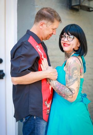 A couple sharing an intimate moment outdoors, with the woman in a turquoise dress showing colorful arm tattoos.