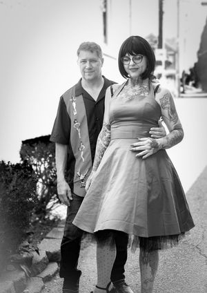Black and white portrait of a couple in casual clothing standing outdoors, with the woman wearing a vintage-style dress and glasses.