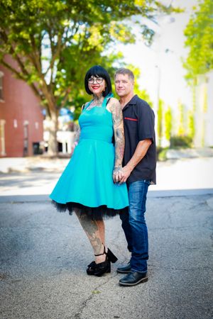 A couple poses outdoors, with woman wearing a bright turquoise dress with black petticoat and the man in casual attire.