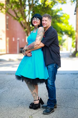 A couple embraces outdoors, with one wearing a bright turquoise dress with tulle and visible tattoos.