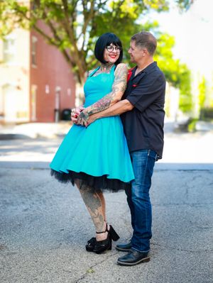 A couple in a romantic pose on a street, with her wearing a vibrant turquoise vintage-style dress and visible tattoos.