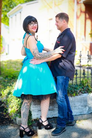 A couple embraces outdoors, with her wearing a bright turquoise vintage dress and showing off intricate leg tattoos.