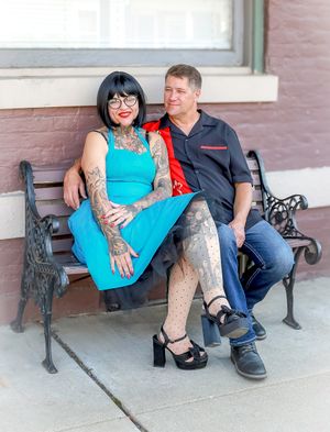 A couple sits together on an ornate metal bench, with the woman wearing a bright turquoise dress and showing tattoo artwork.