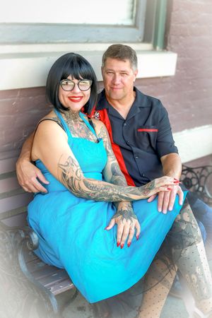 A couple sits together on steps, with a woman in a bright blue dress with tattoos wearing glasses next to a man in dark shirt.