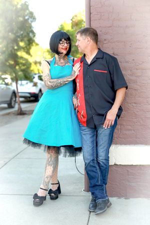 Couple poses against a brick wall, with her wearing a vibrant blue dress and him in casual attire.