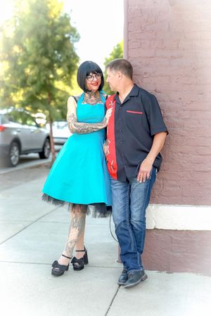 A couple stands against a pink wall outdoors, she wearing a bright turquoise dress with tattoos and he in casual attire.