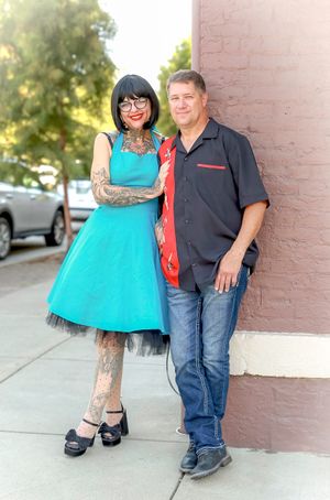 A couple stands against a pink wall, with one wearing a turquoise dress and tattoos while the other wears jeans and dark shirt.