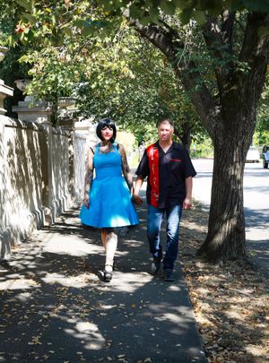 Couple walking together on a tree-lined sidewalk on a sunny day, her in a blue dress and him in casual wear.