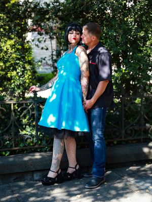 A couple poses outdoors in a garden setting with a bright blue vintage-style dress and casual denim attire.