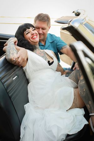 A couple sits in a vintage car sharing laughter and embracing on their wedding day.