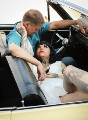 A couple in a vintage convertible car, with the person in white wearing visible tattoos and dark bangs.