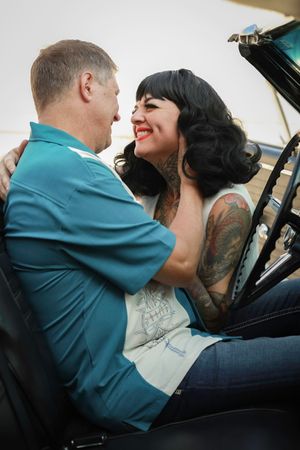 A couple shares a romantic moment leaning on a car door together, with visible tattoos and vintage-inspired styling.