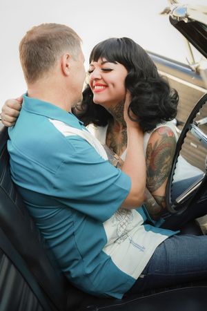 Romantic vintage-style couple embraces in classic car, woman in white dress with tattoos and retro black hairstyle.