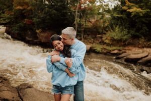 Couple amoureux s’enlaçant tendrement près d’une rivière en forêt, portant des chandails bleus.