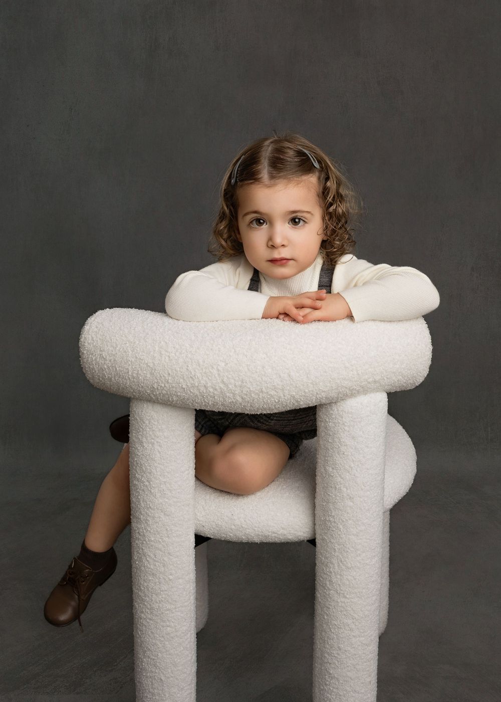 Young child with curly hair poses leaning over a white boucle stool, wearing suspenders, in a studio portrait.
