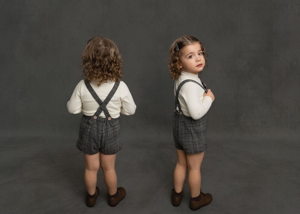 Two toddlers in matching plaid suspender shorts and white shirts pose back-to-back on a gray studio background.