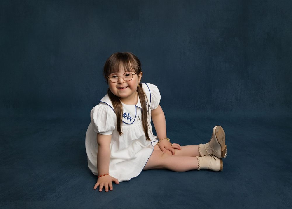 Young girl with Down syndrome in white dress sits smiling on blue studio backdrop for a portrait photo.
