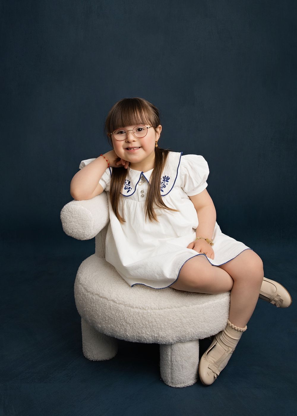 Young girl with glasses in white dress sitting on fluffy chair against dark blue background.
