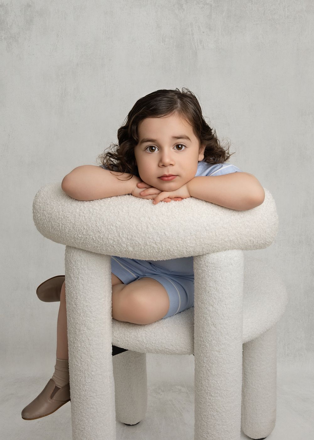 Young child in blue outfit resting chin on arms over a white boucle chair, studio portrait photography.